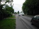 Road with tarmac pavement and grass verge on left.
Large trees on right.
Cars on road.