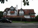 Three-storey house with white-painted walls and variety of red-tiled roofs.
Fence in front of house has an estate-agent's sign.
Grass verge with street-light pole.
Road with markings for a right-turn lane.