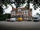 Old Court Hotel.
Red brick building with strong horizontal white features.
Dark tile roofs.
Cars and vans parked around the building.
Overhanging trees.