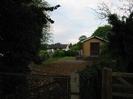 Small service building of sandy-coloured brick.
Fence with white cottage behind.
Dark trees either side.