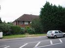 Road with pavement, grass verge, and bank of trees.
Two-storey house of dark red brick with brown tile roof.