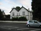 White two-storey house with double pitched roof of dark slate.
Low white wall along road.
Car on road.
Dark trees on right.