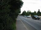 Looking east on the Bath Road.
Bank of trees on the left.
Pavement.
Road with queue of approaching cars.
Low walls with signs on far side of road.
Street lights.