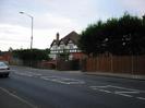 Large house with triple gables above first-floor windows.
Dark red tile roof.
Wooden slat fence alongside road, with conifer trees behind.
Street light.