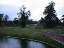 River in foreground.
Sloping river bank with brick wall on right.
Trees with open field beyond and more trees on the horizon.