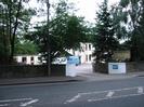 Vehicle exit from Silchester House School.
Low walls along road with white-painted section marking driveway.
Trees partly obscuring two-storey cream-painted building with tall windows.
Lower building on left.
Parked minibusses.
Telephone pole in pavement.