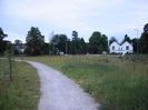 Wide footpath surfaced with grey chippings.
Scrubby areas each side with newly-planted trees.
Wooden railing fence in the distance with road and houses beyond.
White house on right with double-pitch roof.
Street-lights along road.