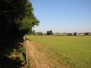 Grassy field on right with farm buildings and houses beyond.
Footpath on left with trees.