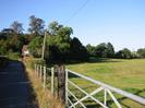 Looking north on Hitcham Road.
Road on left with house in the distance.
Barbed-wire fence and metal gate leading to open field on right.
Mobile-phone mast disguised as a tree on the horizon.