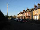 Looking north on Hitcham Road.
Grass verge on left with dark bushes and telephone pole.
Row of houses on the right.
Cars parked in the road outside the houses.