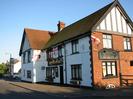The Maypole public house.
White painted walls on the left, with red brick end-wall.
Black window frames.
Several St George's Cross flags flying on the pub.
Arched cellar window low down on the right.