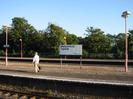 Railway platforms with sign "Welcome to Taplow. Thames Trains"
Red lamp-posts.
Iron railings and large trees beyond far platform.
Passenger walking along platform.