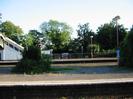 Island platform with bushes and lamp-poles.
Far platform has iron railings dividing it from the car-park beyond.
Large trees in the background.