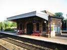 Small station building on island platform.
Red brick with arch details in light stone.
Canopy with decorative edging.
Red metal bench seats.
Notices and timetables.