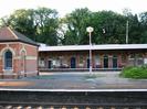 Island platform with small building of red brick with light stone arches and dark slate roof.
Far platform has larger building of similar construction, with long canopy.
Large trees in the background.