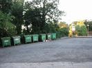 Car park with row of recycling containers.
Trees in the background.
Iron railings and station platform on the right.