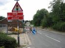 Road with roadworks.
Roadsign warning of traffic lights and new road layout.
Buildings on left.
Trees on right.