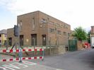 Large brick building with narrow windows and dark wooden doors.
Traffic light.
Gate and entrance to yard on right.
Barriers around roadworks.