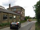 Road with traffic lights and digger.
Buildings on left - partly single storey and partly three-storey.
Trees on right.