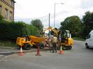 Road junction with digger and dump-truck.
Hedge and building on left.
Telephone pole and street-light.
Trees on right.