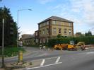 Road junction with traffic lights and bollard on left.
Three-storey building with lower building behind.
Large hedge.
Roadworks with yellow dump-truck and digger.