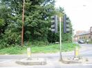 Road junction with traffic lights.
Traffic islands with bollards.
Trees on far side of road with telephone pole in grass verge.
Single-storey building on right with pitched roof and clerestory windows.