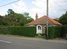 Bungalow with white front door and red pyramid-shaped roof.
Hedges and telephone pole alongside road.
