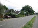 Looking south on Marsh Lane.
Low houses on left, with bushes and trees in front.
Estate agent's sign.
Grass verge and footpath on right.