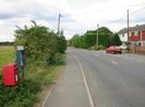 Looking north on Marsh Lane.
Pillar box and parish notice-board on the left.
Hedge, footway and road.
High conifer hedge and houses on the right.