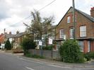 Road with houses on the far side.
Nearest house is set end-on and has three storeys with patterned brickwork.
Estate agents signs.
Bus-stop sign and power pole.