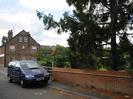 Road with parked car.
Low wall on right with large tree behind.
Three-storey brick building in the background.