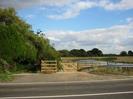 Road in foreground.
Trees, fence, wooden gate and steel barrier.
Jubilee River in the distance.