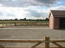 Wooden fence and gate, gravel parking area, small building in patterned brick with tile roof.
Jubilee River in the background.