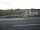 Road with steel barrier and car-park beyond.