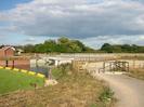 River on left with yellow floating boom and mass of green weed.
Clear water in channel to weir.
Wide path on right surfaced with grey chippings.
Wooden rail fence and black steel kissing-gate.
Buildings and road brifge in the background.