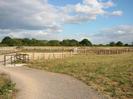 Wide path with grey chippings.
Rough grassy area on right with new trees in containers.
Black steel kissing-gate.
Wooden railings.
Car-park with white height restricting gantry.
Trees in the background.