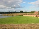 River with mass of green weed held by yellow floating boom.
Small brick building on right.
Trees in background.