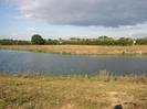 River with scrubby grass on banks.
Trees and houses in distance on far side of river.