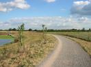 River on left.
Bank slopes up to wide path with grey chipping surface.
Trees in distance on right.