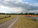 Wide path with field on left.
Slope down to river on right.
Orange mesh enclosures on far side of river, with yellow container on bank.
Trees on horizon.