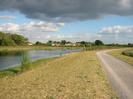 River on left with houses in background.
Scrubby grass bank with wide path on right.