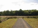 Wide path surfaced with grey chippings.
Scrubby grass on each side.
Wooden fence with black steel kissing-gate at the end, leading to gravel track.
Signpost at junction with road.
Bench on far right.
Bank of trees in the background.