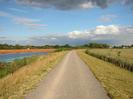 Wide path with river on left and row of newly-planted trees on the right.
Orange mesh enclosures on far side of river.