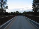 Road across bridge with chevron roadsign at far end.
Bridge has dark wood parapets.