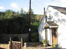 White-painted end wall of bungalow with porch and door.
Telephone pole and railway embankment behind.
Wall and gate in foreground.