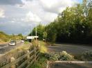 Road and railway embankment on left.
Wooden rail fence.
Car park on right with trees behind.
Tall street-lights.
Cars on road.