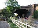 Arch bridge carrying mailine railway across the A4 Bath Road.
Trees on left.
Wooden post-and-rail fence with car-park on left.
Chevron road-sign.