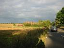Looking east on Approach Road.
Wire fence and open fields on left.
Group of houses set among trees.
Railway embankment on right with large trees.